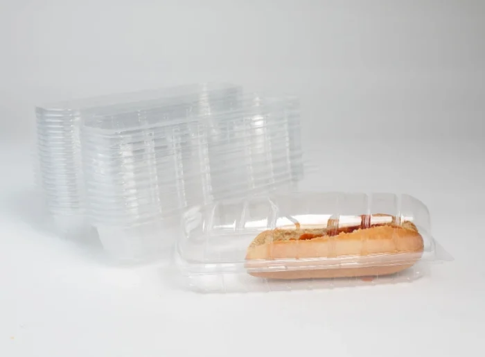 "A stack of transparent hot dog clamshell containers beside a single showcased clamshell with a hot dog, by JC Packaging."