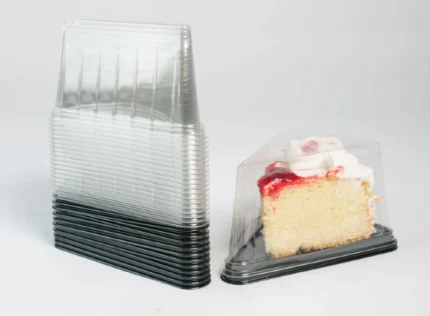 "A stack of JC Packaging's triangular clear cake slice packaging next to a single showcased slice of vanilla cake with whipped cream and strawberry topping, emphasizing the aesthetic and practical design."